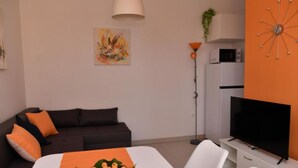 Una sala de estar moderna con un sofá oscuro, una mesa de comedor con un frutero, un televisor de pantalla plana y un armario blanco.
