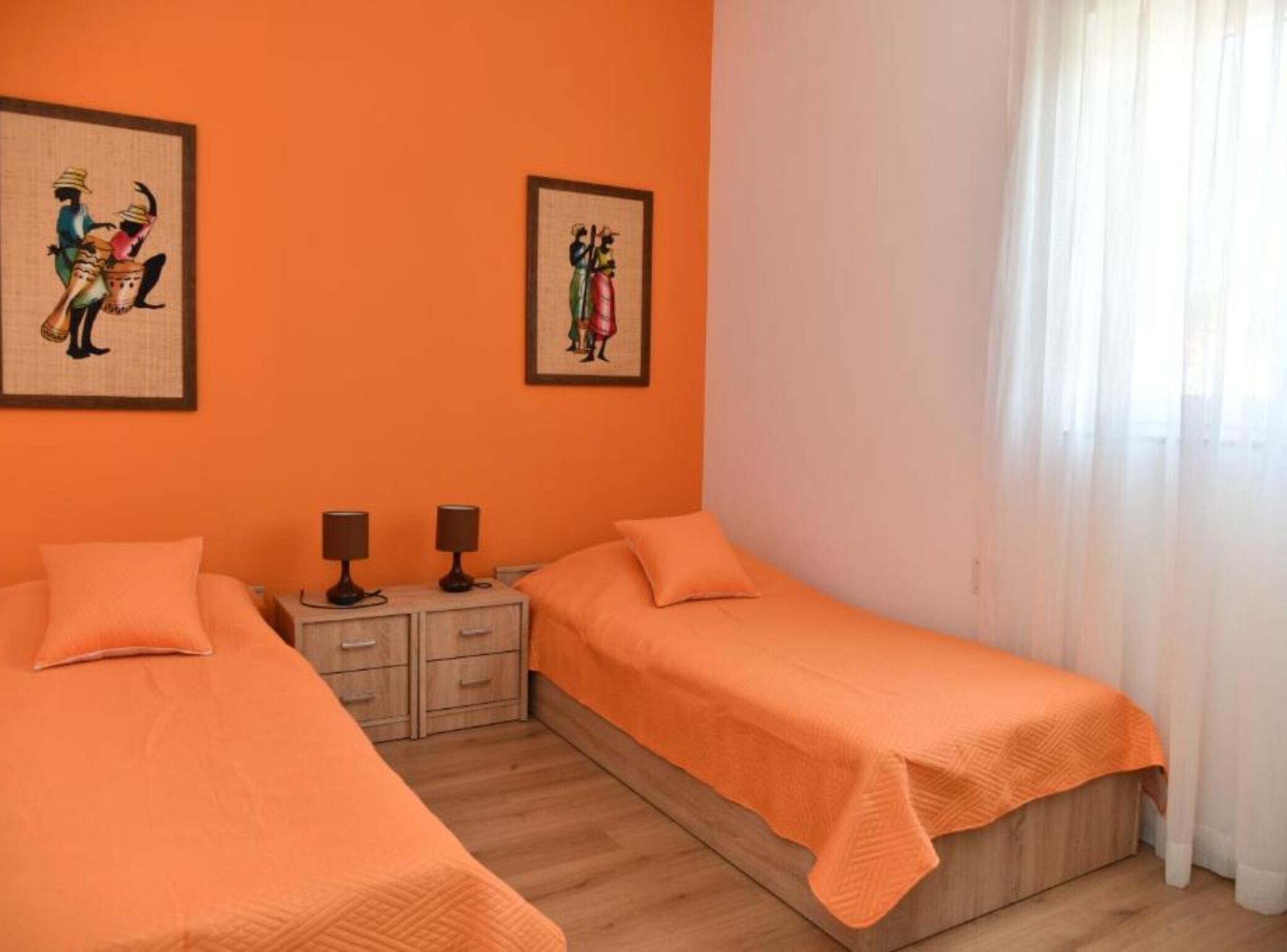 Une chambre avec deux lits, chacun recouvert d’une couverture orange, une table de chevet en bois avec deux lampes, et deux tableaux encadrés accrochés au mur.