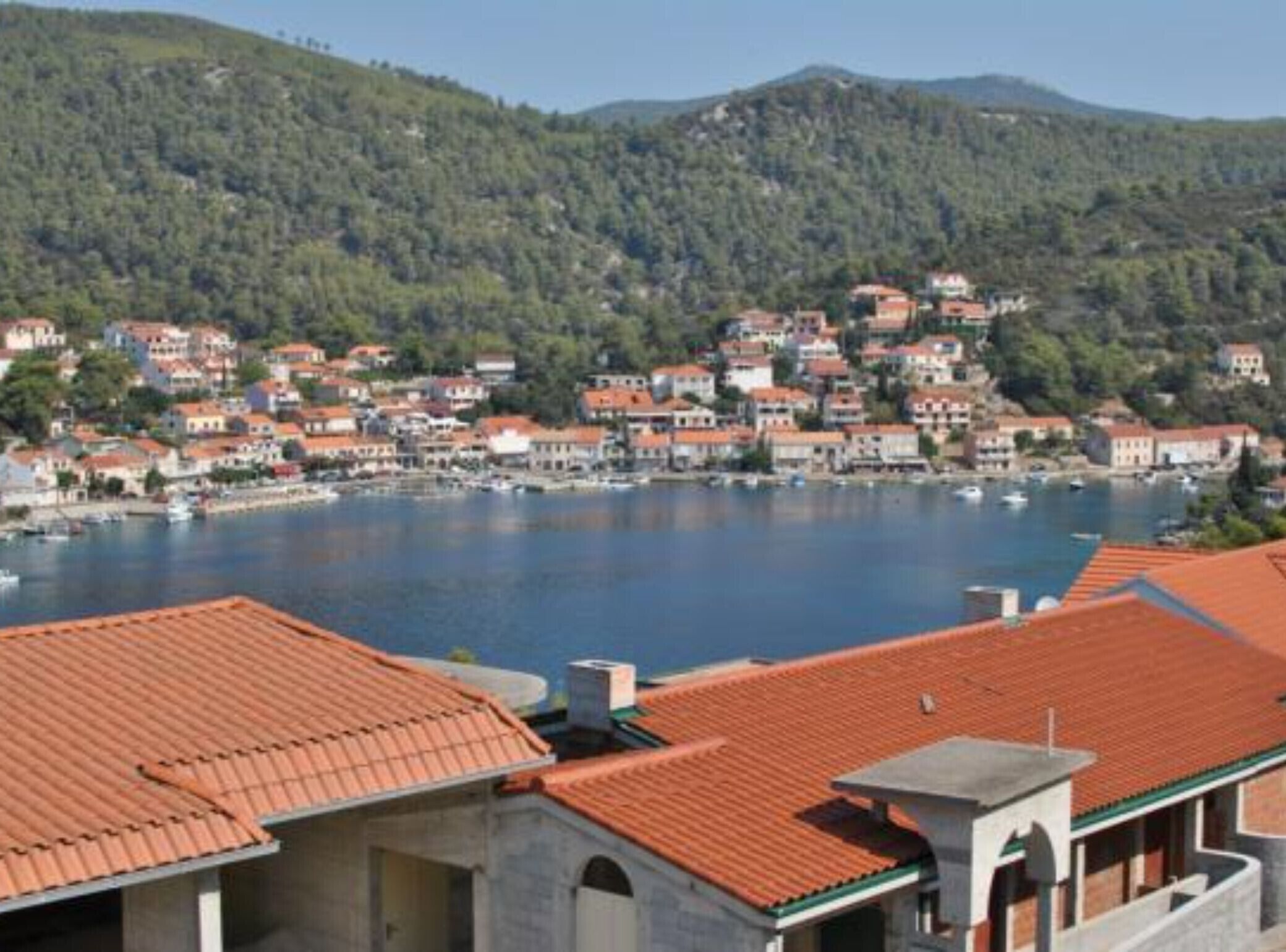 A coastal town with terracotta-roofed buildings, a marina, and hills in the background.