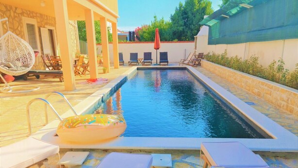 Une piscine entourée de chaises longues et d’un fauteuil suspendu, le tout délimité par un mur de pierre et complété par un espace abrité avec des chaises et une table.