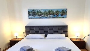 Un lit double bien fait, avec une tête de lit capitonnée, deux oreillers blancs et deux serviettes pliées posées sur le lit. Il y a deux tables de chevet avec des lampes, et un tableau représentant des palmiers est accroché au mur au-dessus du lit.