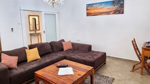 Un salon avec un canapé d’angle marron foncé, une table basse en bois, un lustre, un tableau représentant un coucher de soleil et une table à manger en bois avec des chaises.