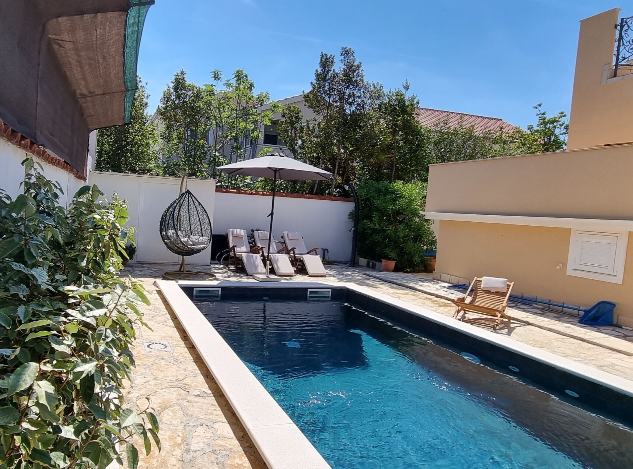 Piscina com espreguiçadeiras e cadeira suspensa, cercada por vegetação e uma parede branca.