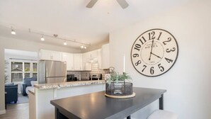 Une cuisine moderne avec des meubles blancs, une grande horloge et un coin repas comprenant une table et des chaises aux couleurs sombres.