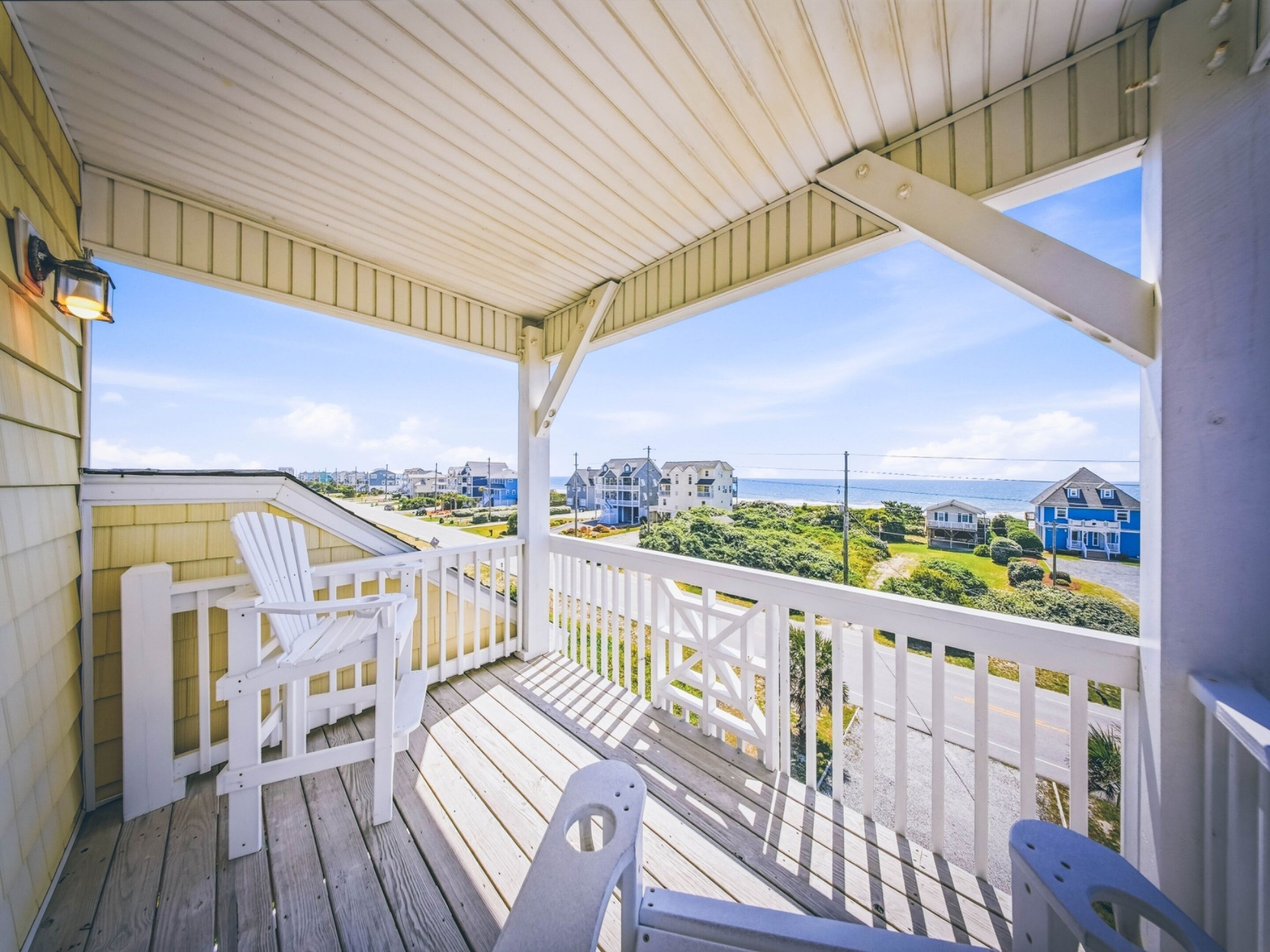 A wooden deck with white railings, a white chair, and a view of houses and the ocean.