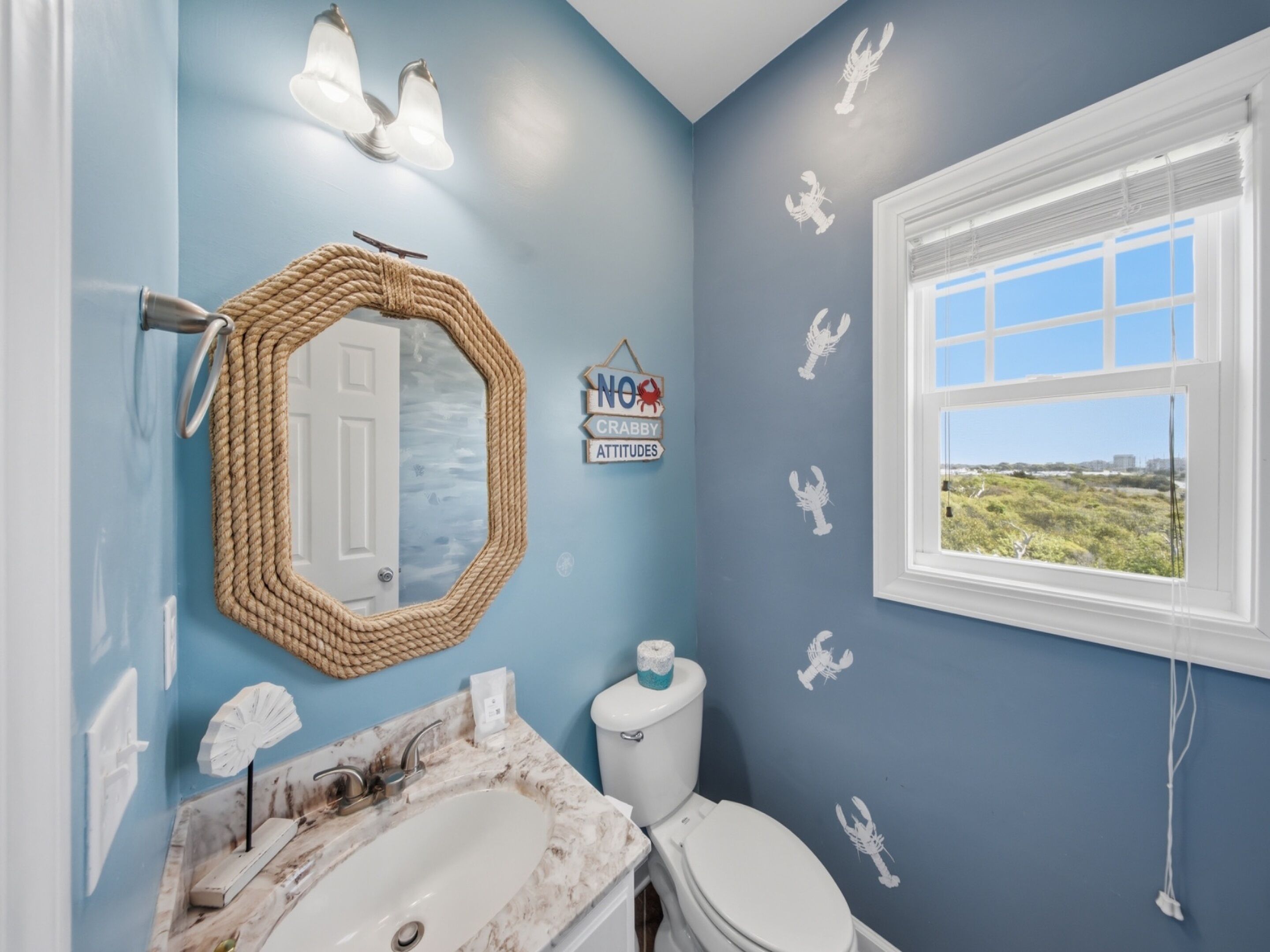 Un bagno con una parete blu, un water bianco, un lavabo con piano in marmo, uno specchio rotondo e una finestra vista verde.