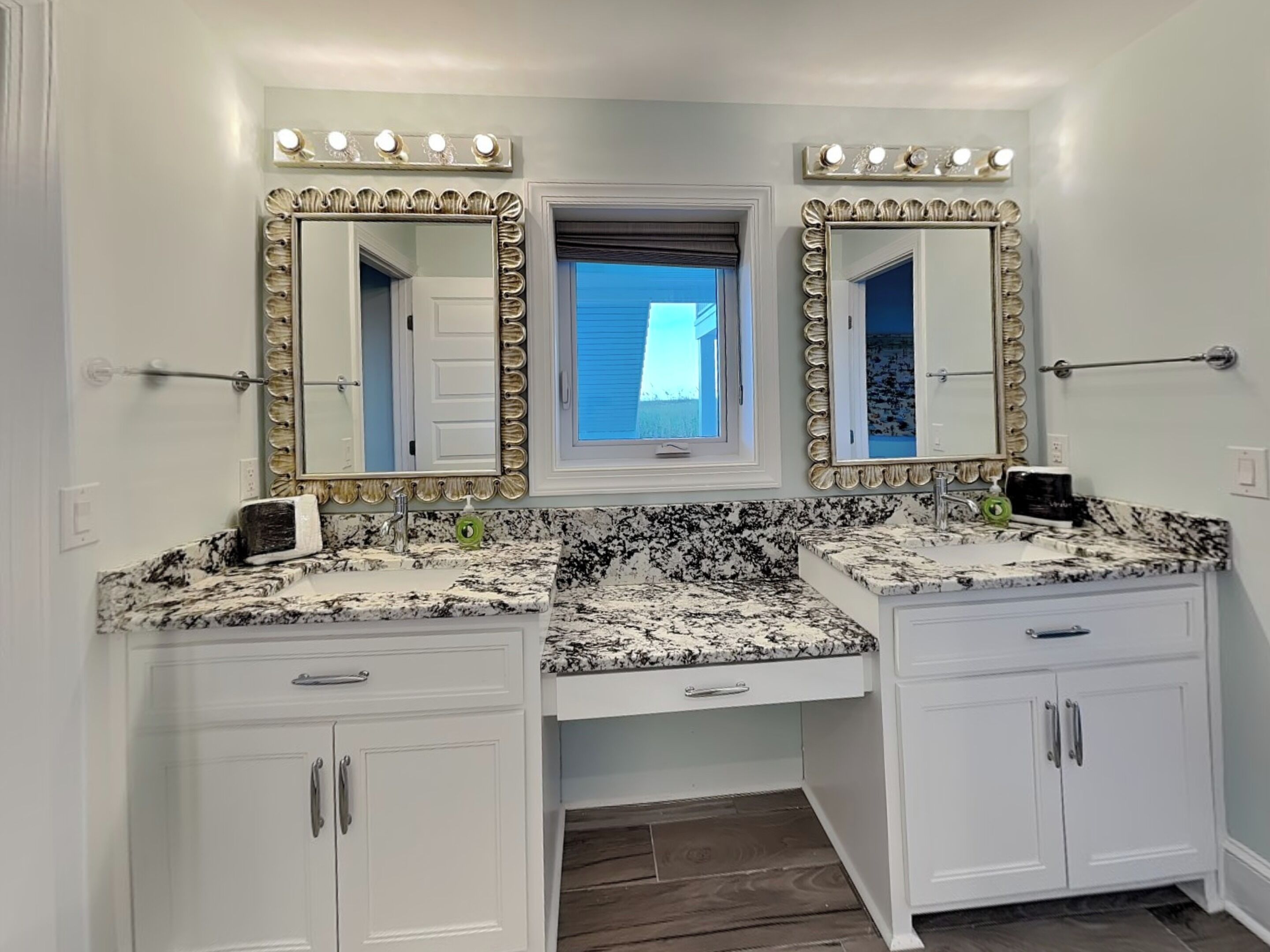 A bathroom with two sinks, a vanity with drawers, a window with blinds, and a mirror with lights above.