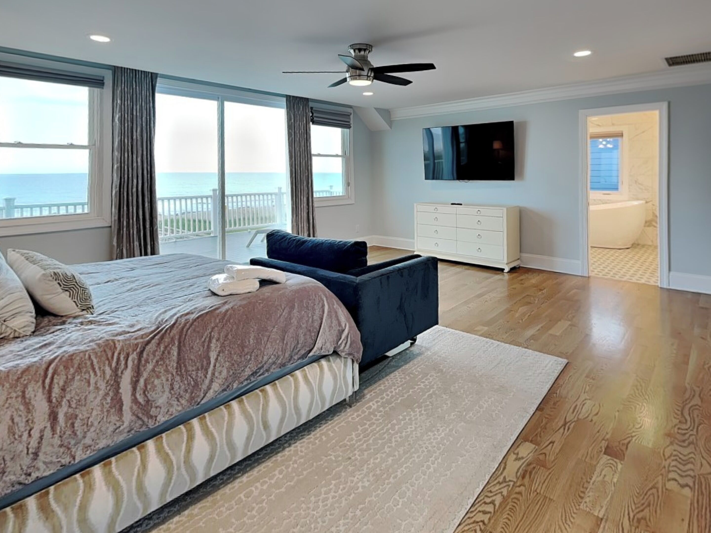 A hotel room with a large bed, a flat-screen TV, a ceiling fan, and a view of the ocean.