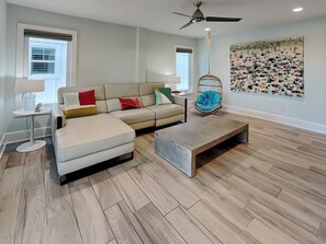 A modern living room with a sectional sofa, a hanging chair, a coffee table, and a wall art piece. - Southern Comfort by Carolina Retreats (Kure Beach)