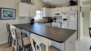 A modern kitchen with white cabinets, a granite countertop, and a central island with bar stools. - Song Of The Sea by Topsail Realty (Holly Ridge)