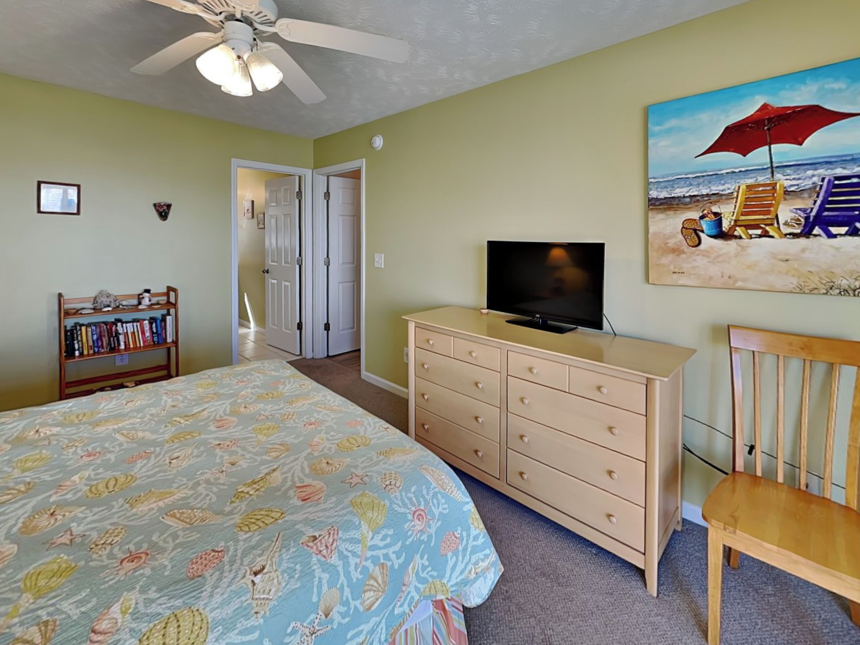 A bedroom with a bed, dresser, television, bookshelf, and a painting of a beach scene.