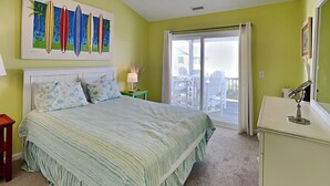 A bedroom with a large bed, a nightstand, a dresser, a ceiling fan, and a view of the balcony through the window. - Song Of The Sea by Topsail Realty (Holly Ridge)