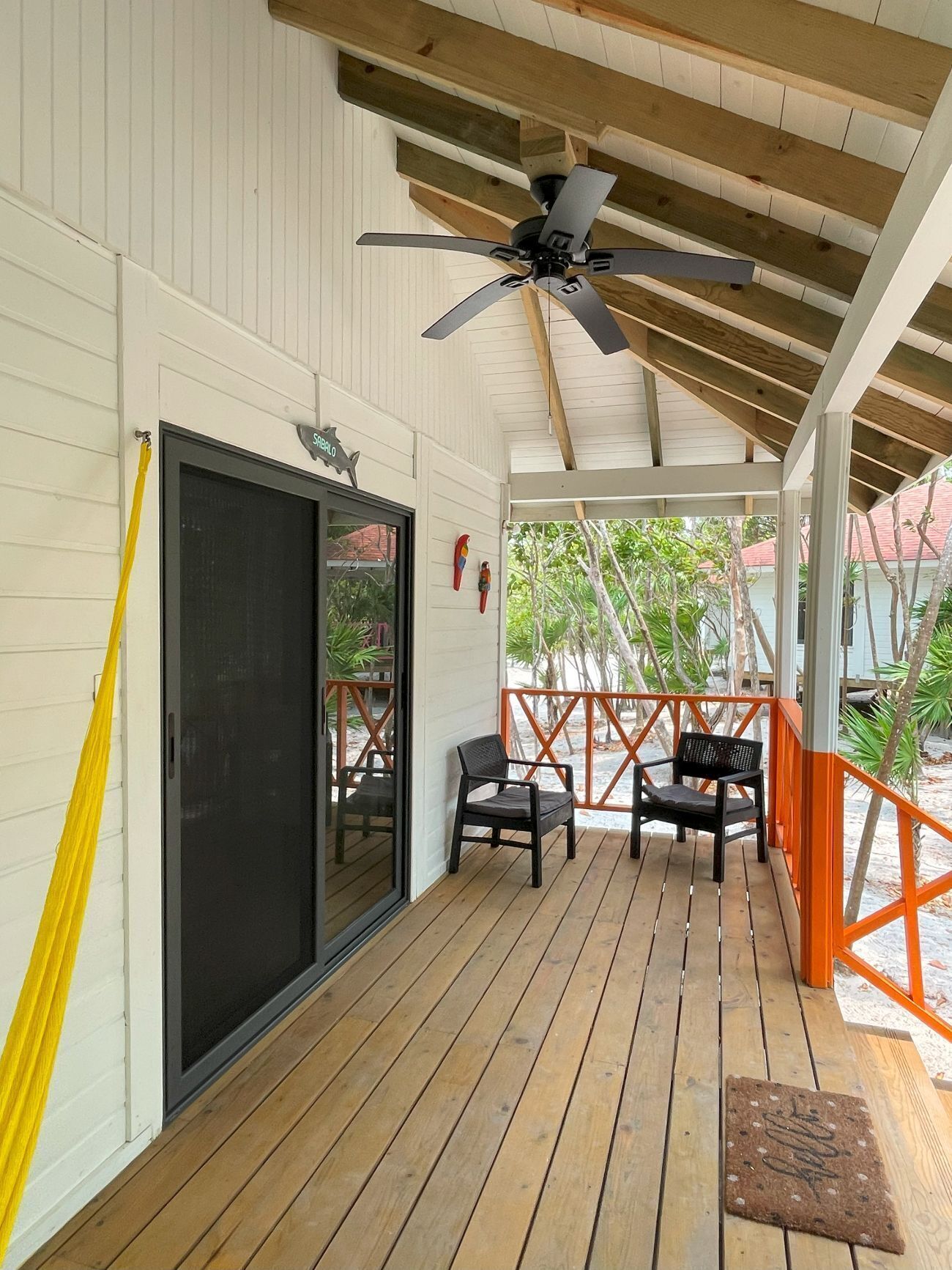 Un espacio exterior techado con suelo de madera, dos sillas, un ventilador de techo y una puerta con mosquitera.
