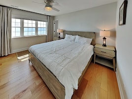 Un dormitorio con una cama grande, suelo de madera, un ventilador de techo, una mesita de noche con una lámpara y una ventana con cortinas.