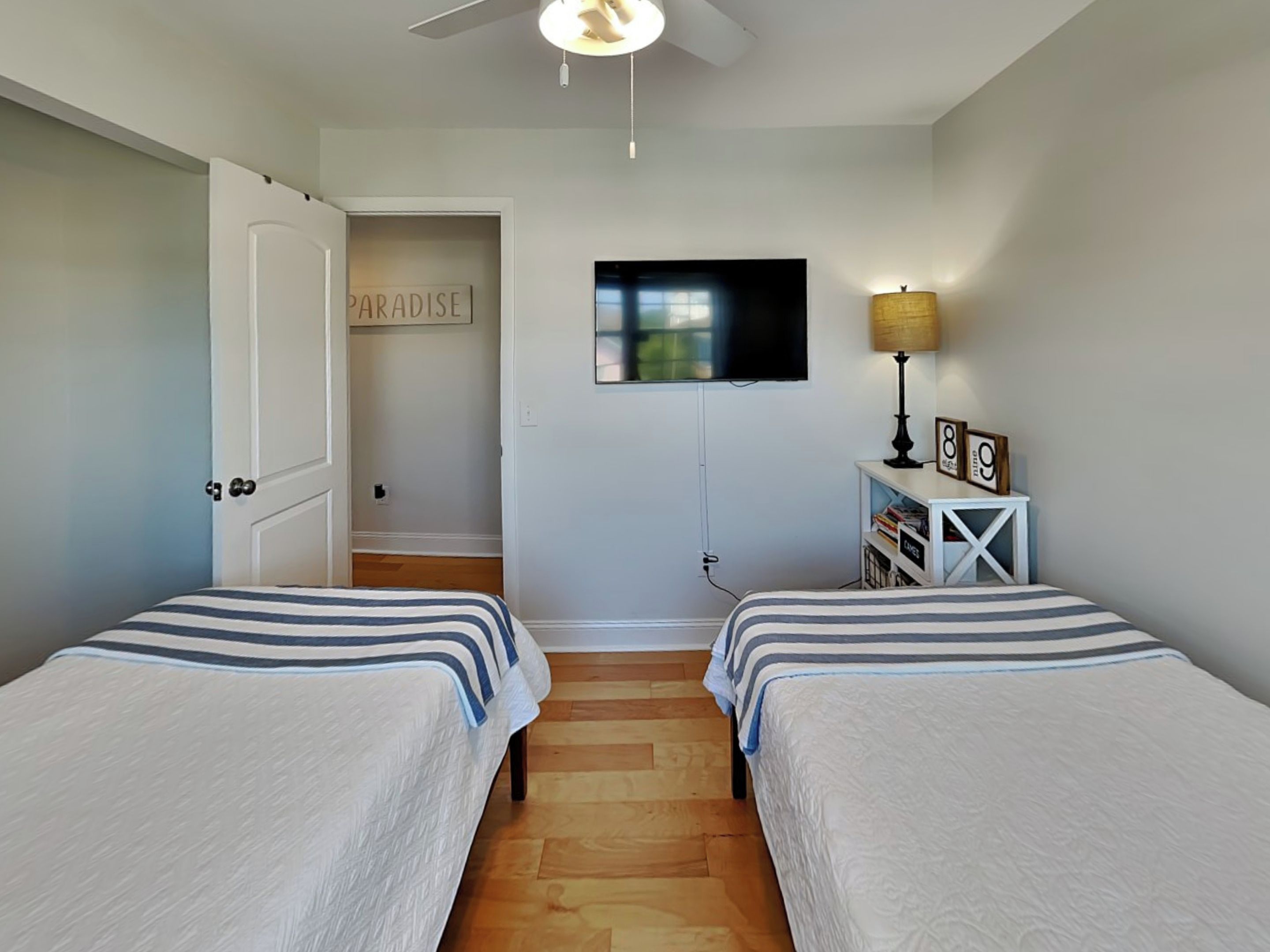 Une chambre avec deux lits, un téléviseur fixé au mur, une porte sur laquelle est affiché un panneau indiquant « PARADIS », et une petite table avec une lampe et des cadres photo.