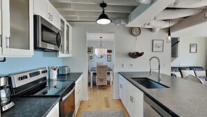 A modern kitchen with stainless steel appliances, a large island with a sink, and a dining area with a patterned rug. - Riptide by Topsail Realty (Holly Ridge)