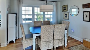 A dining area with a blue table and wicker chairs, a hanging light fixture, and a view through large windows. - Riptide by Topsail Realty (Holly Ridge)