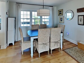 A dining area with a blue table and wicker chairs, a hanging light fixture, and a view through large windows. - Riptide by Topsail Realty (Holly Ridge)