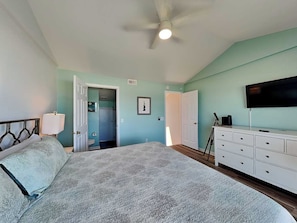 A bedroom with a bed, a ceiling fan, a TV, a dresser, and a bathroom door. - Beauregard's Rest by Carolina Retreats (Carolina Beach)