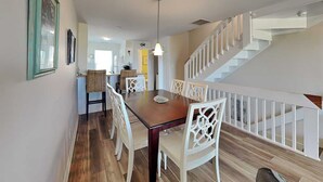 A dining area with a wooden table and white chairs, a staircase with white railings, and a hanging light fixture.