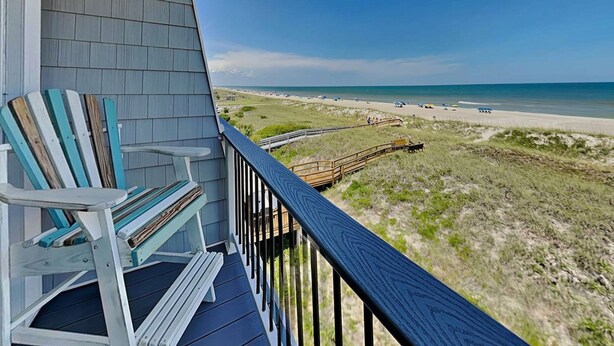A balcony with a view of a beach, a wooden deck, and a chair.