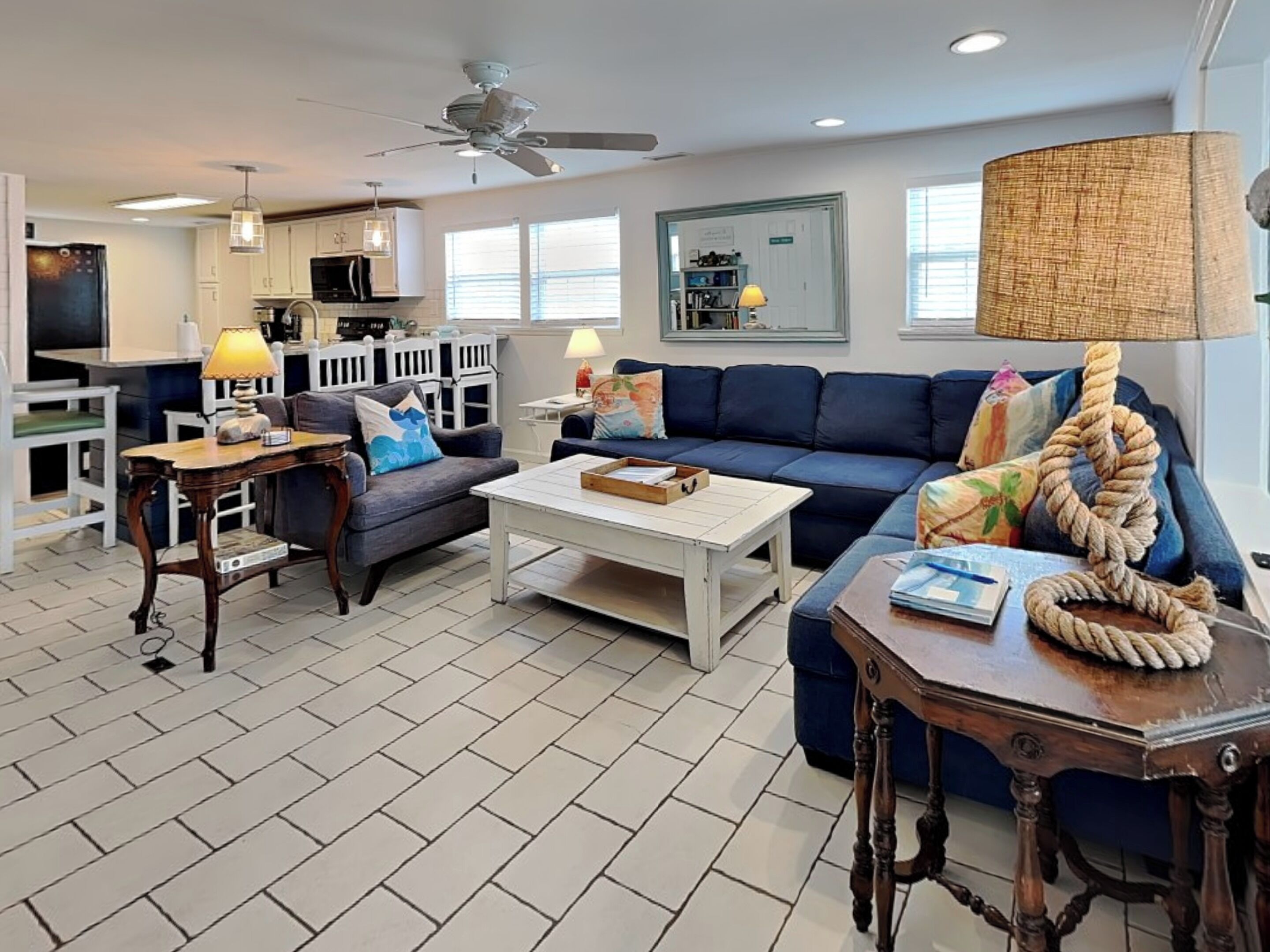 A modern living room with a dark blue sofa, a white coffee table, and a wooden side table. There is a ceiling fan, a lamp, and a large window providing natural light.