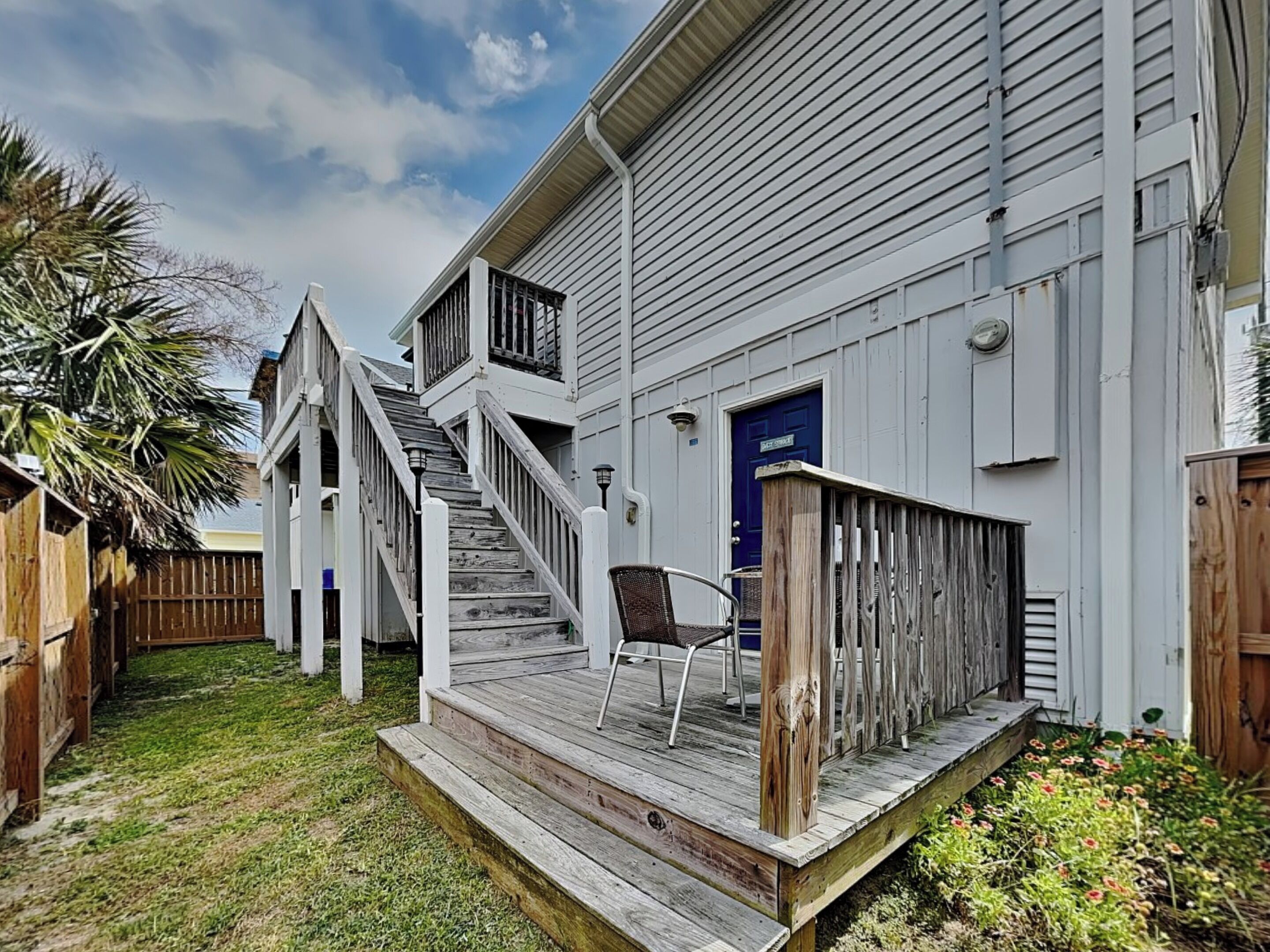 A white house with a wooden deck, a blue door, and a staircase leading to a porch.