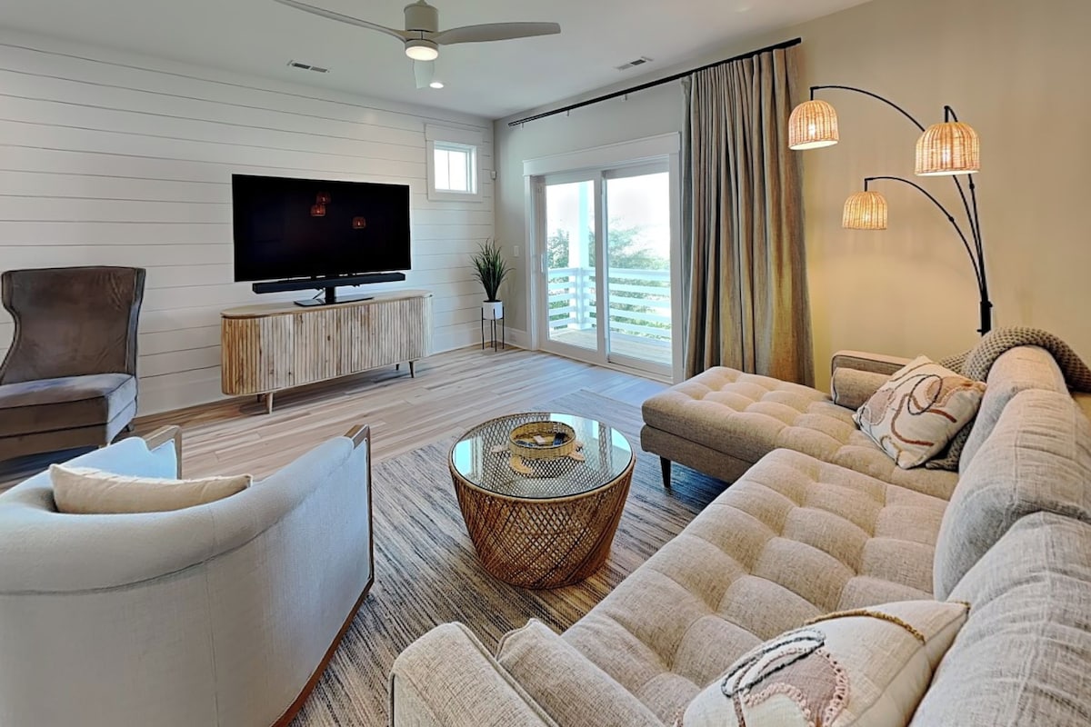 A modern living room with a flat-screen TV, comfortable seating, and large windows.