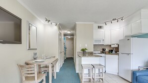 Une cuisine compacte avec des meubles blancs, un four à micro-ondes et un réfrigérateur. On y trouve un coin repas avec une table et des chaises, ainsi qu'un sol à motifs bleus.