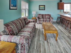 Una sala de estar con un sofá estampado, una mesa de centro de madera y una cocina con taburetes de bar.