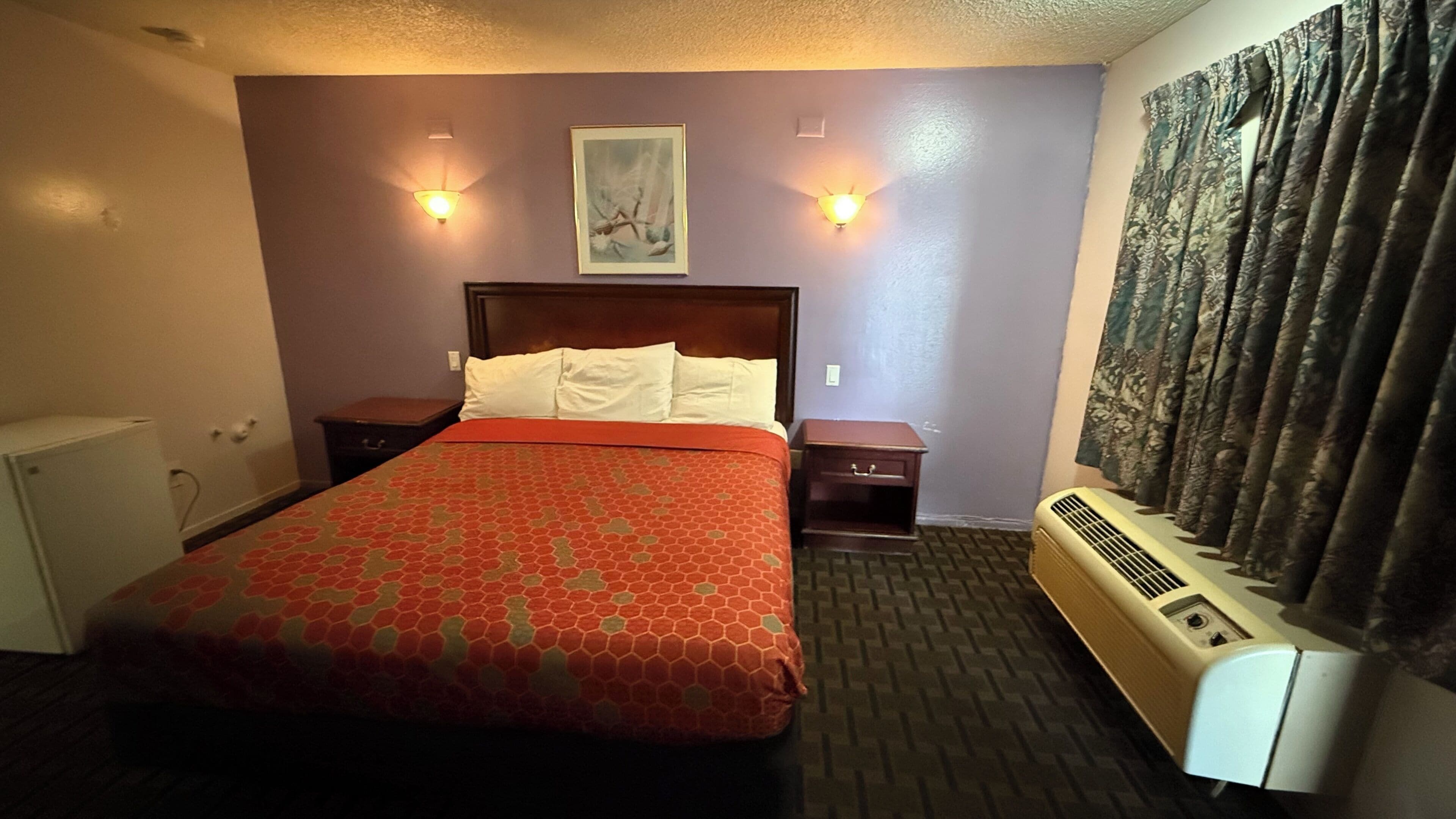 Habitación de hotel con cama, dos mesitas de noche, un calentador, un cuadro en la pared y una ventana con cortinas.