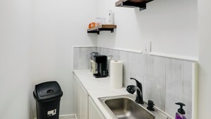 Una pequeña cocina americana con un lavamanos, una cafetera, un cubo de basura y una estantería con un plato y una botella.
