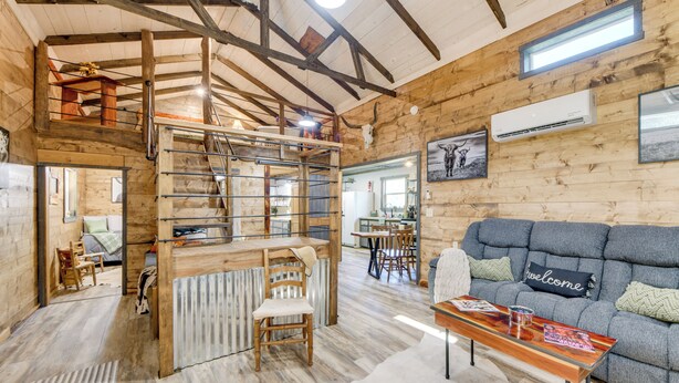 A rustic cabin interior with a loft bedroom, a dining area, and a living room with a sofa and a coffee table. - Lake Eufaula Access: Cozy Wood Cabin w/ Yard! (Abbeville)
