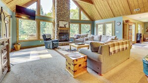 A spacious living room with a stone fireplace, large windows, and a high ceiling.