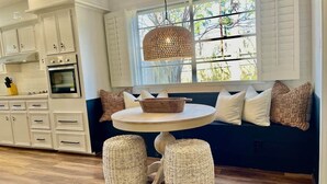 A kitchen with white cabinets, a round table, wicker stools, and a window seat with cushions. - Retreat @ The One | Movie Theater | Coffee Bar (Conroe)