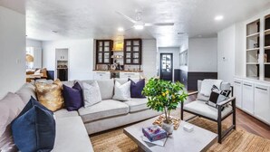 A living room with a sofa, armchairs, a coffee table, a ceiling fan, and a kitchen in the background. - Retreat @ The One | Movie Theater | Coffee Bar (Conroe)