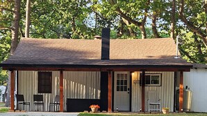 A rustic cabin with a chimney, a porch with chairs, and a welcome sign. - Rustic-Chic 2 bdr Pet Friendly Log Cabin, Steps to Lake Huron (Au Gres)