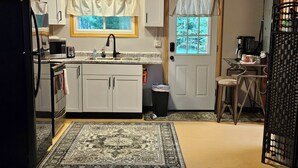 A compact kitchen with white cabinets, a black refrigerator, a microwave, and a coffee maker. There's a window with curtains, a door with a window, and a patterned rug on the floor. - Rustic-Chic 2 bdr Pet Friendly Log Cabin, Steps to Lake Huron (Au Gres)
