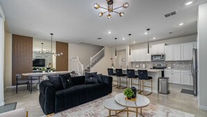 Una sala de estar moderna con un sofá negro, un comedor y una cocina con armarios blancos y electrodomésticos de acero inoxidable.