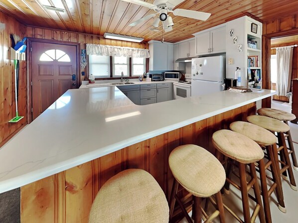 A kitchen with a large island, bar stools, wooden cabinets, and a ceiling fan. - Pana-Sea-A by Topsail Realty (Holly Ridge)