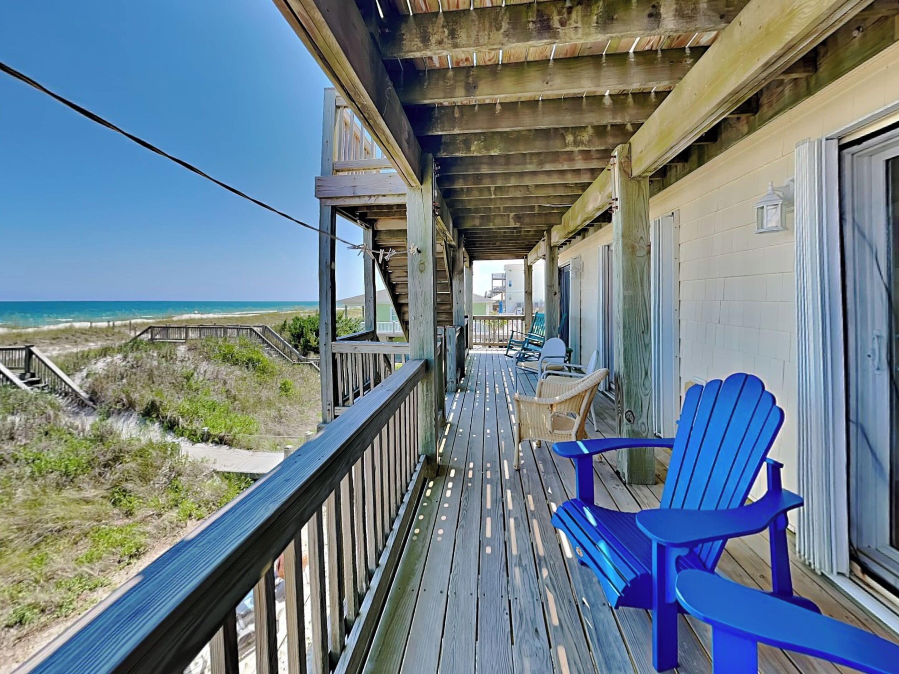 A wooden deck with a view of the beach, a covered area with chairs, and a building with a window.