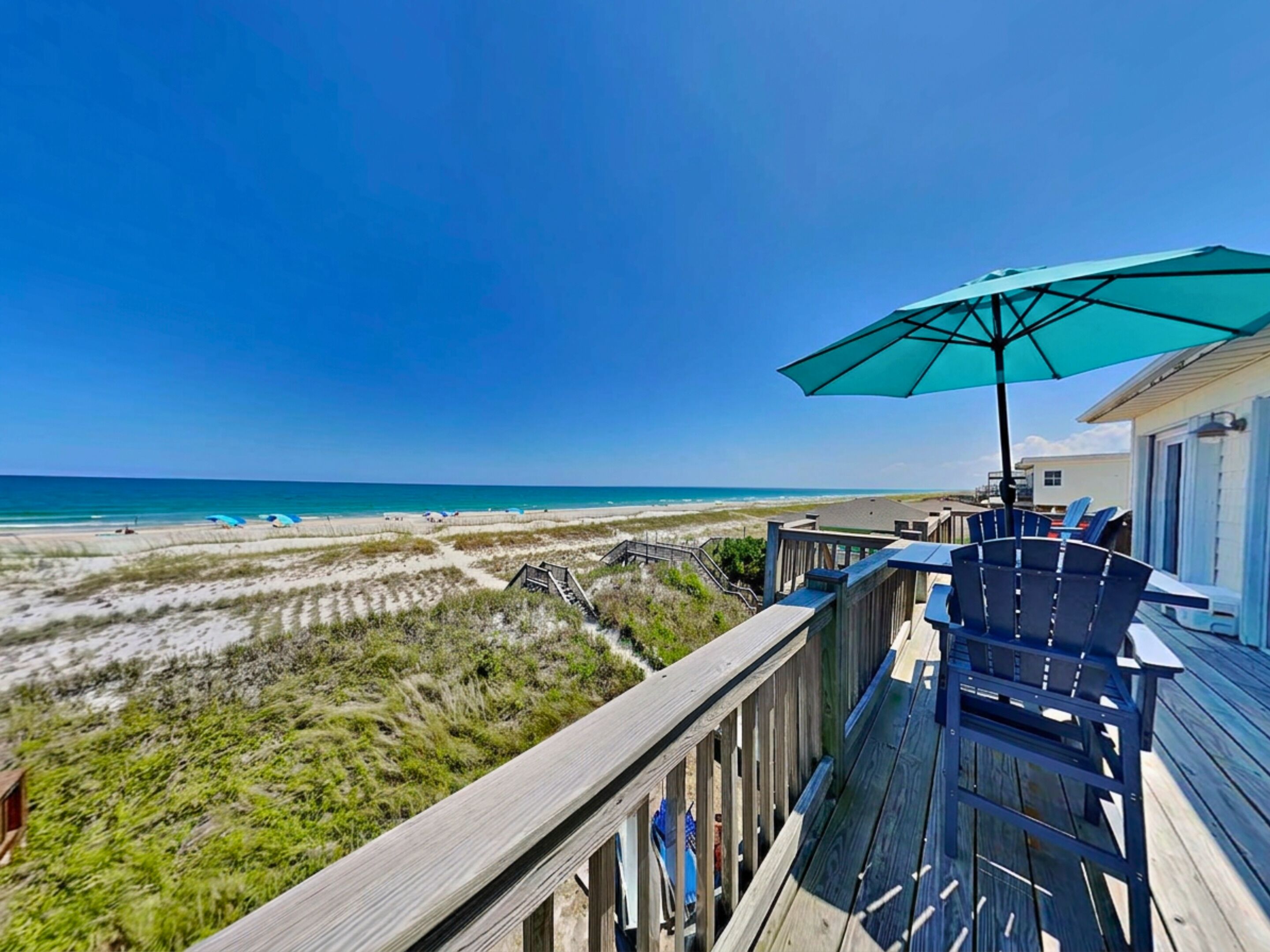 Terraza de madera con mobiliario exterior y una sombrilla grande con vista a la playa.