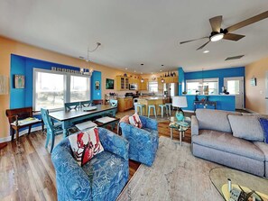 A spacious living area with a dining table, blue patterned sofas, a grey couch, and a kitchen with wooden cabinets. - Piccolo Paradiso by Topsail Realty (Holly Ridge)