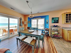 A dining area with a view of the ocean, a kitchen with a blue accent wall, and a dining table with chairs. - Piccolo Paradiso by Topsail Realty (Holly Ridge)