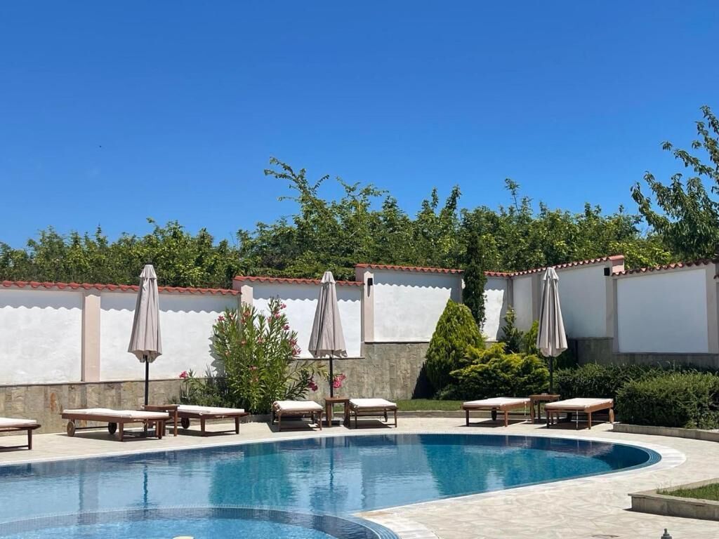 A swimming pool with lounge chairs and umbrellas, surrounded by a white wall and greenery.