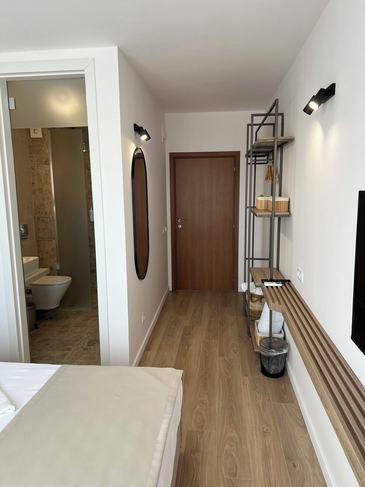 A hotel room with a wooden floor, a wooden door, a small shelf with toiletries, a trash bin, and a wall-mounted light fixture.