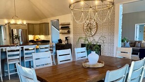 A spacious dining area with a wooden table and white chairs, a chandelier, and a kitchen in the background.