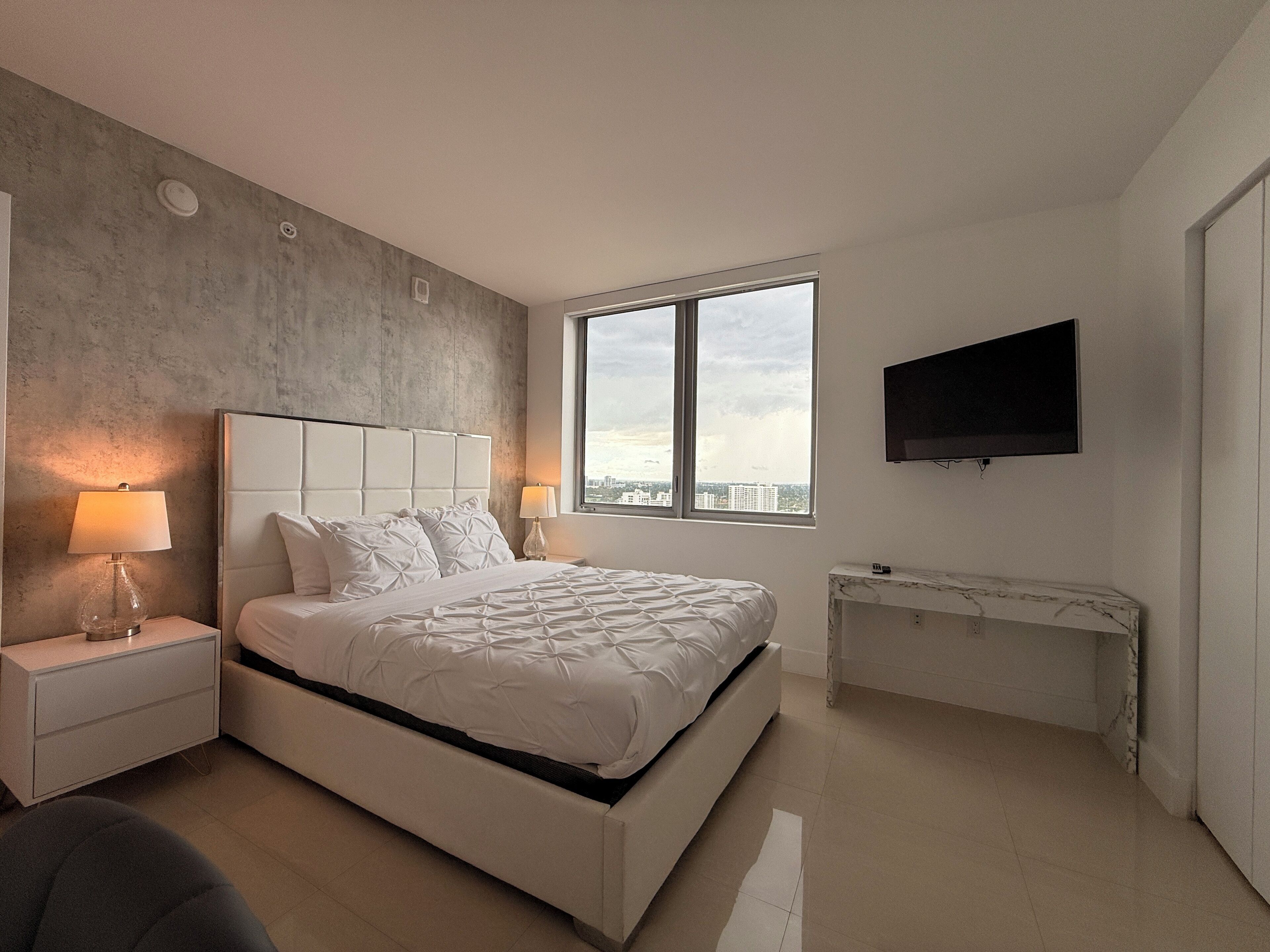 Une chambre moderne avec un grand lit, un téléviseur fixé au mur et une vue sur la ville par la fenêtre.