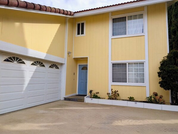 A two-story yellow house with a white garage door, a blue door, and a small front garden.