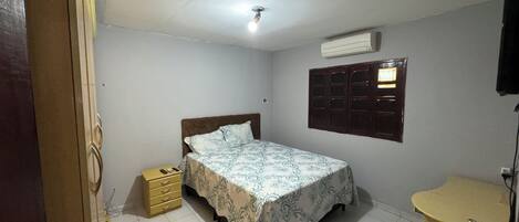 Un dormitorio con cama, mesita de noche, cómoda, ventana con persianas y aire acondicionado de pared.